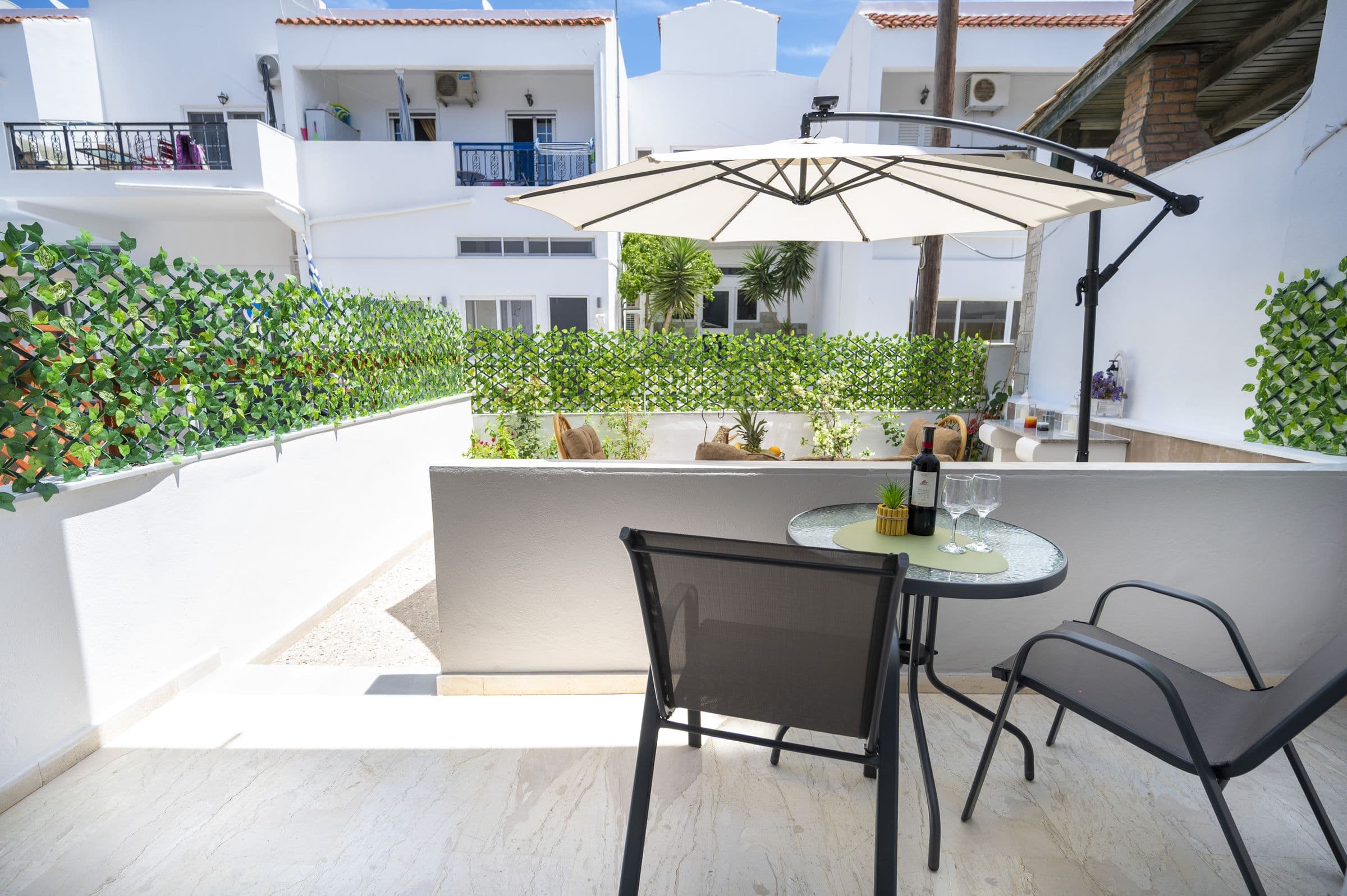 Terrazza con tavolino tondo in vetro e due sedie, affacciata sul cortile con piante e ombrellone